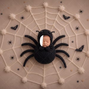 Könnte beinhalten: Ein schwarzes Strickkostüm als Spinne mit Kapuze, Fühlern und acht Plüschbeinen, getragen von einem Säugling. Das Kostüm ist mittig auf einem großen, cremeweißen Häkel-Spinnennetz platziert, das mit kleinen schwarzen Spinnen, Fledermäusen und winzigen Sternen verziert ist.