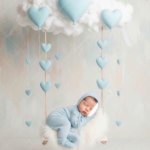Op de afbeelding: Een slapende pasgeborene in een lichtblauwe gebreide outfit, rustend op een zacht wit oppervlak. Boven hangt een wolkvormige decoratie met blauwe hartvormige ballonnen, met kleinere harten eronder. De scène creëert een dromerige, grillige sfeer.