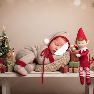 Puede incluir: Un bebé dormido con un traje de elfo beige y rojo descansa sobre una almohada junto a una muñeca elfo navideña. Un pequeño árbol de Navidad, una figura de reno y regalos envueltos están en un banco blanco.