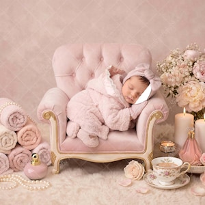 May include: A sleeping baby in a pink outfit and matching bonnet rests on a small, ornate pink chair. The scene includes rolled towels, candles, flowers, and perfume bottles, creating a soft, feminine aesthetic.