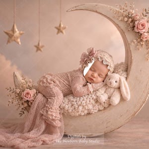 Pode incluir: Um recém-nascido com uma touca de renda e um macacão de manga comprida a condizer, a dormir num grande acessório em forma de meia-lua cor de creme decorado com rosas cor-de-rosa. Um pequeno coelho de peluche está sentado ao lado do bebé.