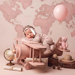 May include: A newborn baby in a pilot's hat and outfit rests on a pink wooden airplane prop. A pink teddy bear, a globe, a suitcase, and a balloon complete the travel-themed nursery setup. The background features a world map.