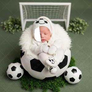 May include: A newborn baby in a white soccer-themed outfit, including a hat, mittens, and booties, nestled inside a soccer ball. The scene is set on a green surface with a miniature soccer goal and additional soccer balls.