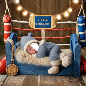 May include: A newborn baby wearing a blue knitted outfit and hat, lying on a faux fur bed inside a miniature boxing ring. The ring has red ropes, blue and red punching bags, and a sign that reads "Little Knockout".