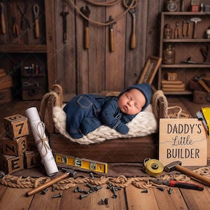 Può includere: Un neonato in tuta blu dorme in un lettino di legno, circondato da attrezzi e blocchi di legno. Un cartello recita "Daddy's Little Builder". La scena è ambientata in un'officina rustica.