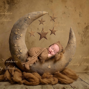 May include: A newborn baby sleeping on a crescent moon-shaped prop decorated with stars. The baby is wearing a brown knitted outfit and is surrounded by a brown fuzzy blanket. The background is a textured brown wall with the text "The Newborn Baby Studio" written in cursive.
