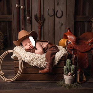 May include: A baby dressed in a cowboy outfit, including a hat and boots, lies on a fluffy white blanket inside a wooden crate. A brown leather saddle and a coiled rope are nearby, with a cactus in a pot. The scene is set in a rustic, wooden-themed environment.