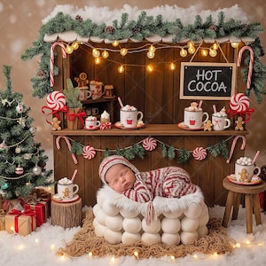 Puede incluir: Un bebé recién nacido con un atuendo a rayas rojas y blancas duerme en una cama redonda blanca frente a un puesto de chocolate caliente en miniatura con temática navideña. El puesto tiene un letrero que dice "HOT COCOA" y está decorado con bastones de caramelo, hombres de jengibre y un pequeño árbol de Navidad.