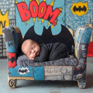 May include: A sleeping baby in a black outfit rests on a gray fur-like surface inside a small Batman-themed chair. The chair has a black bat-shaped back and is set against a backdrop with comic book-style graphics, including the word "BOOM".