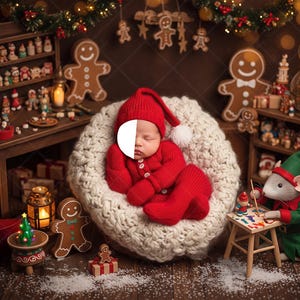 May include: A newborn baby wearing a red knit outfit and hat with a white pom-pom, nestled in a cream-colored knitted nest. The Christmas-themed scene includes gingerbread men, miniature toys, and decorative lights.