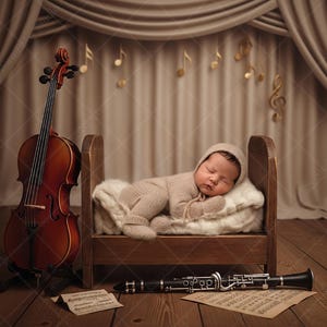 Könnte beinhalten: Ein Neugeborenes in beiger Kleidung und Mütze schläft in einem kleinen Holzbett. Eine Cello steht links, eine Klarinette und Notenblätter unten. Noten hängen am Vorhang.