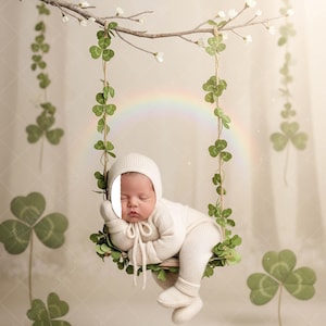 Può includere: Un neonato con un completo e cappuccio in maglia bianca, dorme pacificamente su un'altalena di legno adornata con foglie verdi e trifogli. Un arcobaleno sullo sfondo crea una scena serena e stravagante.