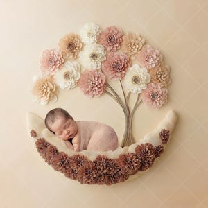 May include: A newborn baby sleeps peacefully in a crescent-shaped prop decorated with brown flowers. Above, a floral tree with pink, white, and beige paper flowers creates a soft, dreamy scene. The background is a light beige with a subtle diamond pattern.