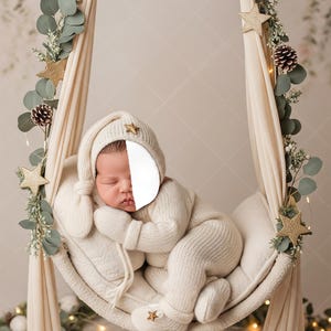 May include: A newborn baby wearing a cream-colored knit outfit, including a hat, mittens, and booties, rests in a cream-colored hammock. The hammock is decorated with greenery, gold stars, and pinecones. The background is a soft, neutral color.