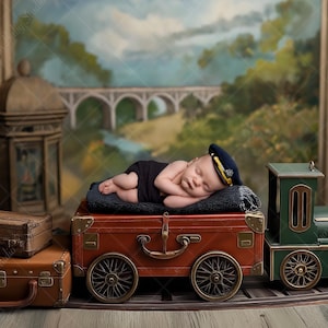 May include: A newborn baby wearing a navy blue conductor hat and black outfit, sleeping on a red vintage suitcase. A green toy train is next to the suitcase. The background features a painted landscape with a bridge and trees.