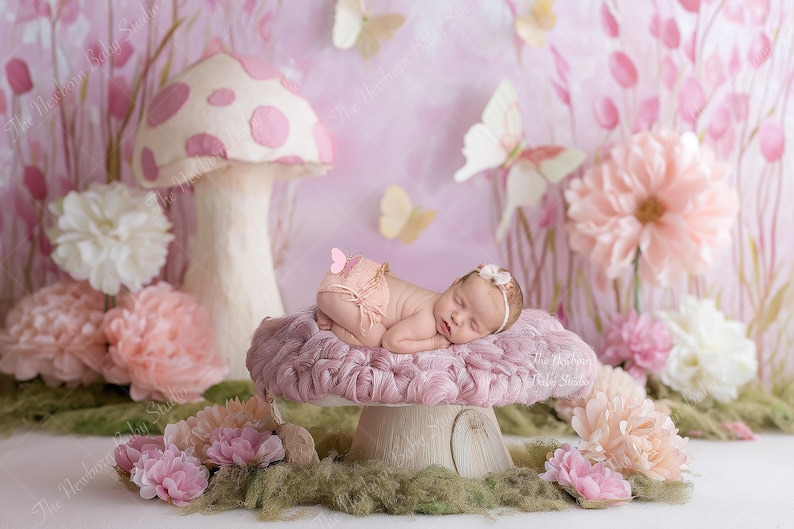 May include: A newborn baby lies peacefully on a plush, pink mushroom prop, surrounded by floral decorations and butterflies. The backdrop features a soft pink and white color scheme, creating a whimsical and dreamy scene for newborn photography.