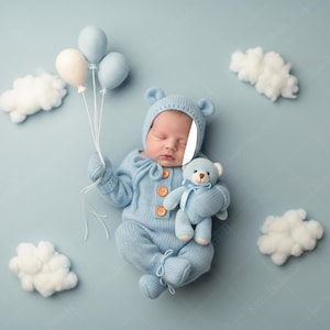 May include: A newborn baby wearing a light blue bear-themed outfit, including a hat with ears, and holding a matching teddy bear and balloons. The baby is lying on a light blue surface with white cloud decorations.