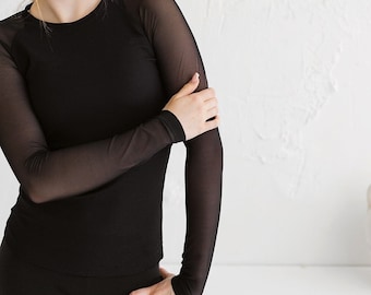 Puede incluir: Una mujer con una camisa negra de manga larga con una superposición de malla transparente. La camisa tiene una textura acanalada y un escote redondo.