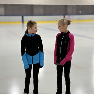 Könnte beinhalten: Zwei junge Mädchen in schwarzen Jacken mit blauen und rosa Akzenten stehen auf einer Eisbahn. Beide tragen schwarze Hosen und weiße Schlittschuhe. Das Mädchen links hat ihre Haare zu einem Pferdeschwanz gebunden, während das Mädchen rechts ihre Haare zu einem Dutt gebunden hat. Beide schauen sich an und lächeln.
