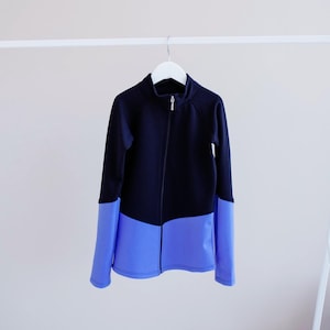 Puede incluir: Una chaqueta negra y azul con cremallera y cuello alto. La chaqueta tiene la parte superior negra y la inferior azul.