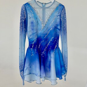 K&ouml;nnte beinhalten: Ein blaues und wei&szlig;es Eiskunstlaufkleid mit langen &Auml;rmeln und einem mehrlagigen Rock. Das Kleid ist mit Strasssteinen verziert.