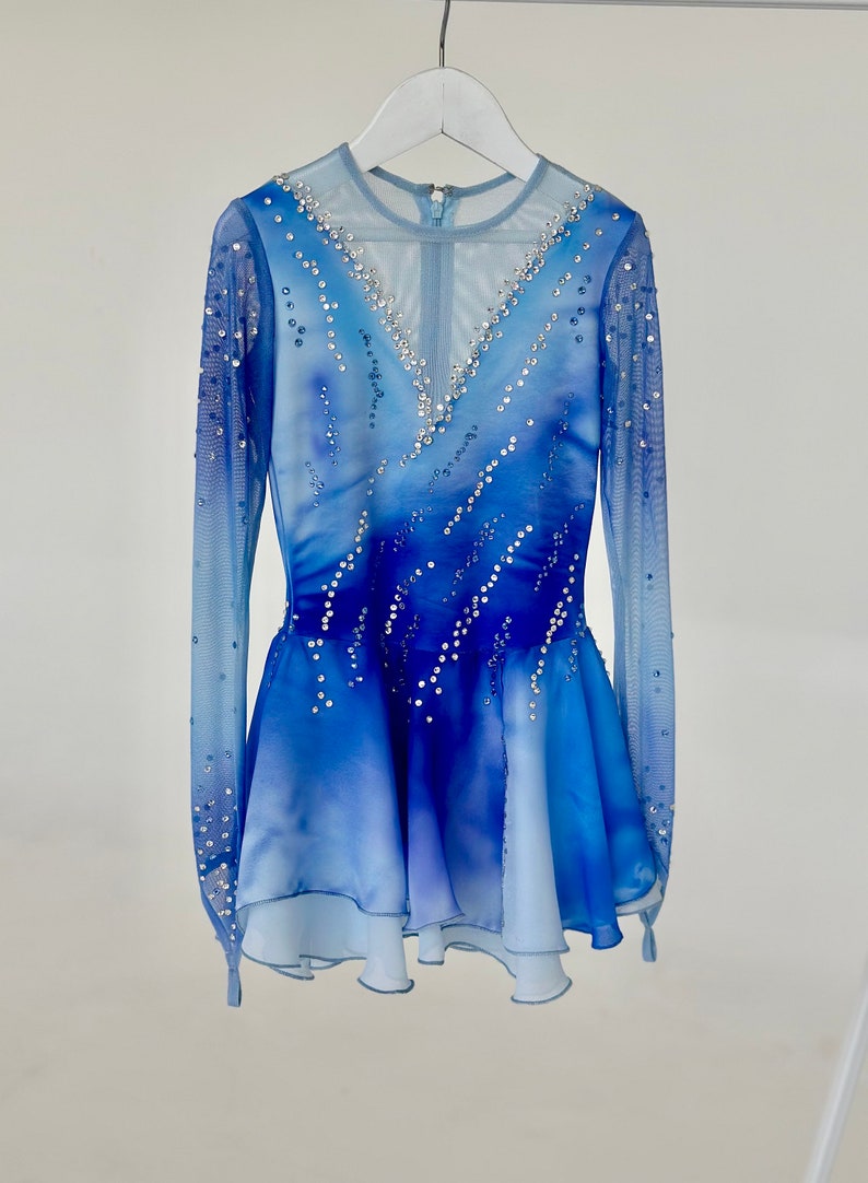 K&ouml;nnte beinhalten: Ein blaues und wei&szlig;es Eiskunstlaufkleid mit langen &Auml;rmeln und einem mehrlagigen Rock. Das Kleid ist mit Strasssteinen verziert.
