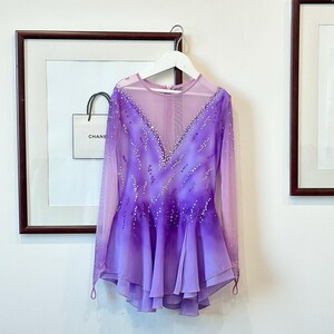 May include: A purple and pink figure skating dress with long sleeves and a mesh bodice. The dress is decorated with rhinestones and has a layered skirt.