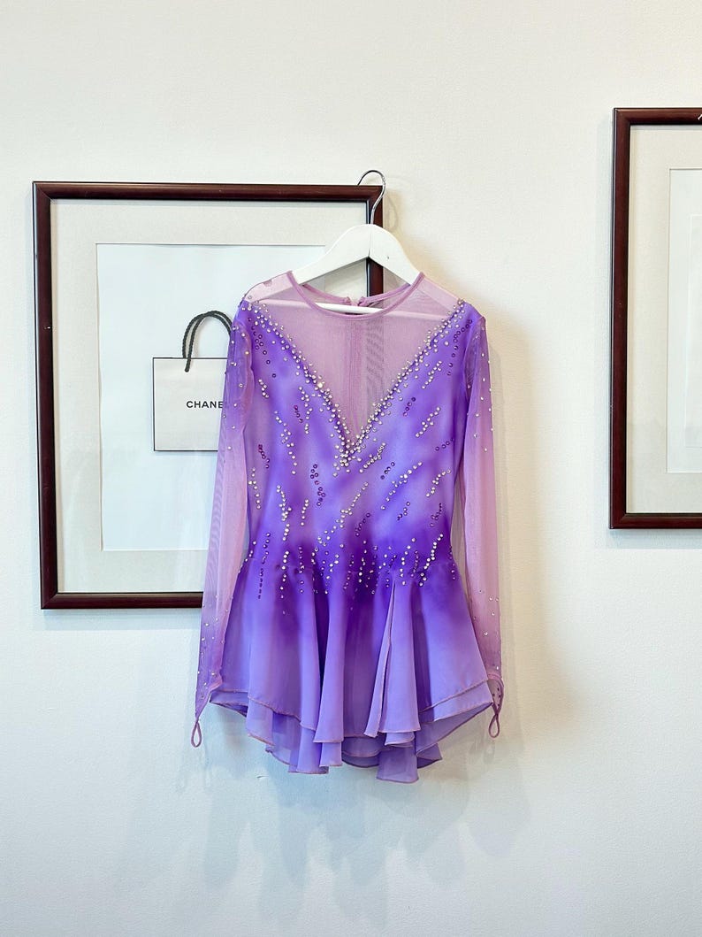 May include: A purple and pink figure skating dress with long sleeves and a mesh bodice. The dress is decorated with rhinestones and has a layered skirt.