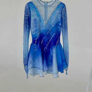 K&ouml;nnte beinhalten: Ein blaues und wei&szlig;es Eiskunstlaufkleid mit einem durchsichtigen, lang&auml;rmeligen Oberteil und einem mehrlagigen Rock. Das Kleid ist mit Strasssteinen verziert.