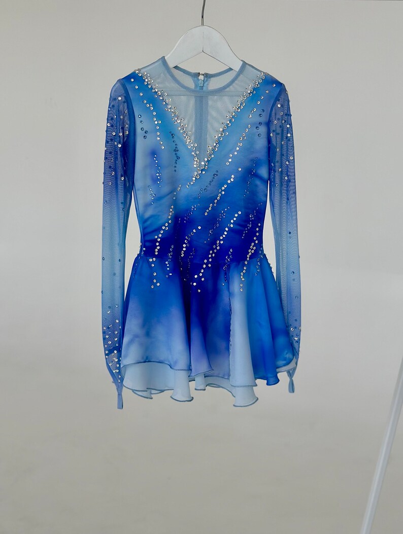 K&ouml;nnte beinhalten: Ein blaues und wei&szlig;es Eiskunstlaufkleid mit einem durchsichtigen, lang&auml;rmeligen Oberteil und einem mehrlagigen Rock. Das Kleid ist mit Strasssteinen verziert.