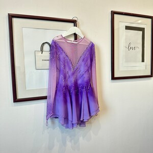 May include: A purple and pink figure skating dress with long sleeves and a sheer bodice. The dress is decorated with rhinestones and has a gradient design.