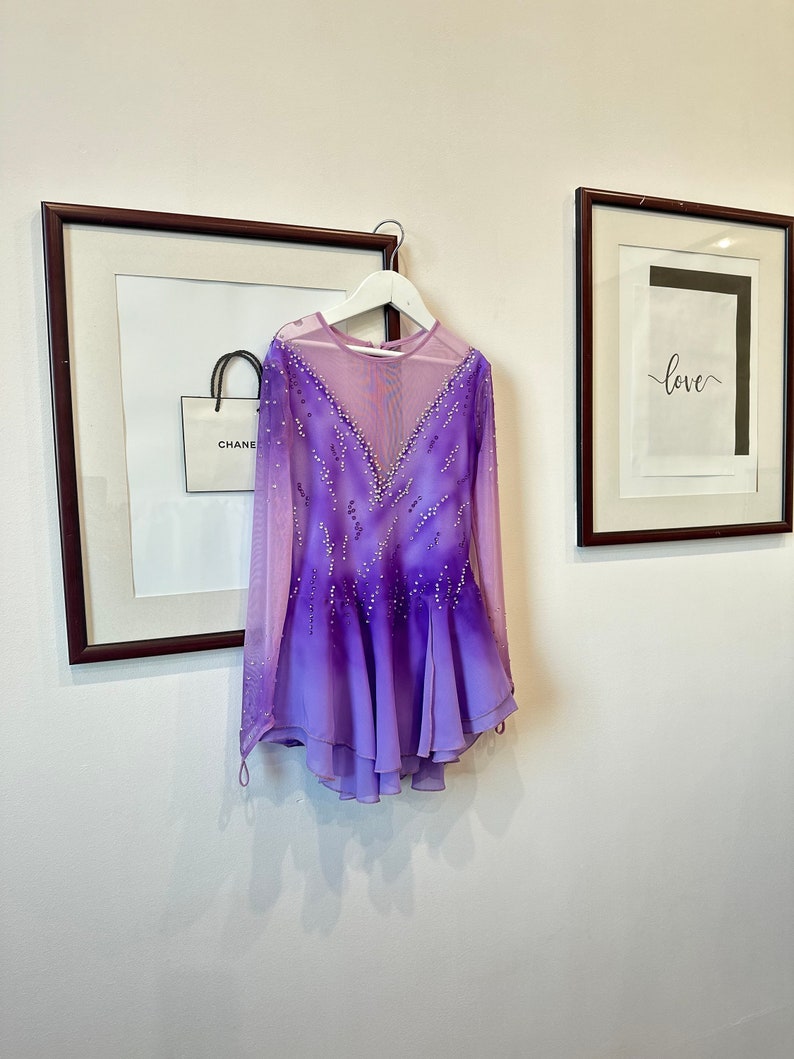 May include: A purple and pink figure skating dress with long sleeves and a sheer bodice. The dress is decorated with rhinestones and has a gradient design.