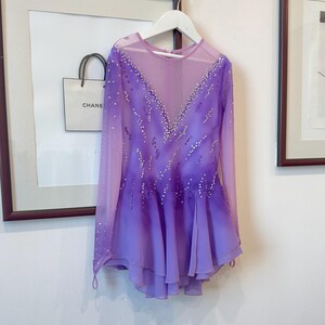 May include: A purple figure skating dress with long sleeves and a sheer bodice. The dress is decorated with rhinestones and has a gradient from a darker purple at the top to a lighter purple at the bottom.