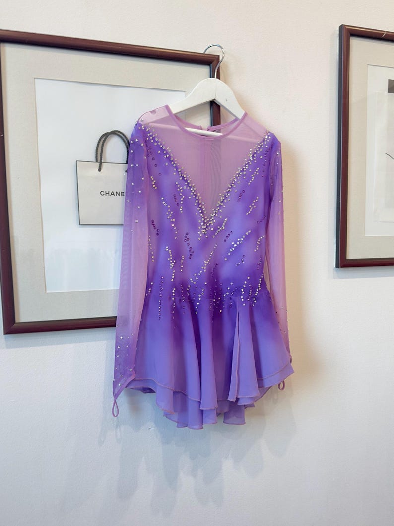 May include: A purple figure skating dress with long sleeves and a sheer bodice. The dress is decorated with rhinestones and has a gradient from a darker purple at the top to a lighter purple at the bottom.