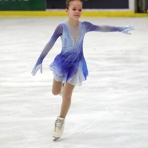 K&ouml;nnte beinhalten: Eine junge Eiskunstl&auml;uferin in einem blau-wei&szlig; gemusterten Kleid mit wei&szlig;em Rock und wei&szlig;en Schlittschuhen. Die L&auml;uferin f&uuml;hrt einen Pirouettensprung auf dem Eis aus.