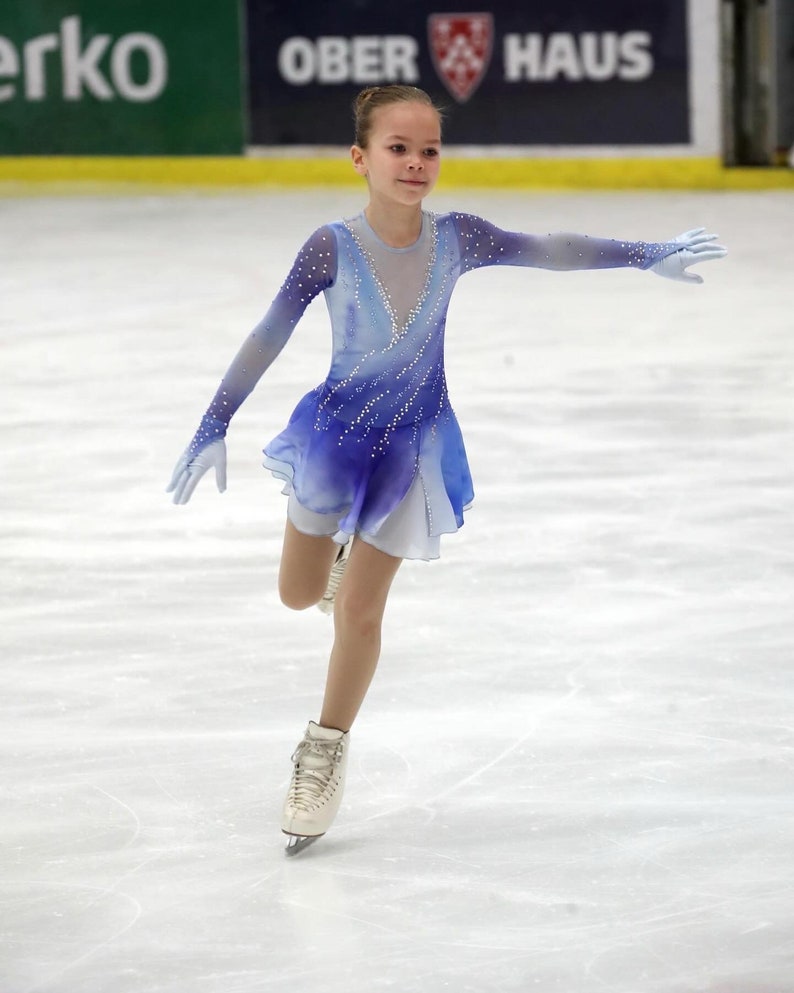 K&ouml;nnte beinhalten: Eine junge Eiskunstl&auml;uferin in einem blau-wei&szlig; gemusterten Kleid mit wei&szlig;em Rock und wei&szlig;en Schlittschuhen. Die L&auml;uferin f&uuml;hrt einen Pirouettensprung auf dem Eis aus.