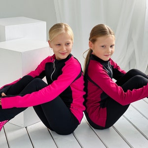 May include: Two girls wearing black leggings and pink and black zip-up jackets. The jackets have black accents and the text "A-GO" on the left side. The girls are sitting on a white wooden floor.