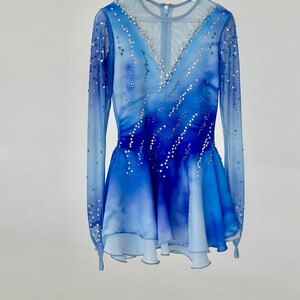 K&ouml;nnte beinhalten: Ein blaues und wei&szlig;es Eiskunstlaufkleid mit langen &Auml;rmeln und einem V-Ausschnitt. Das Kleid ist mit Strasssteinen verziert und hat einen mehrlagigen Rock.