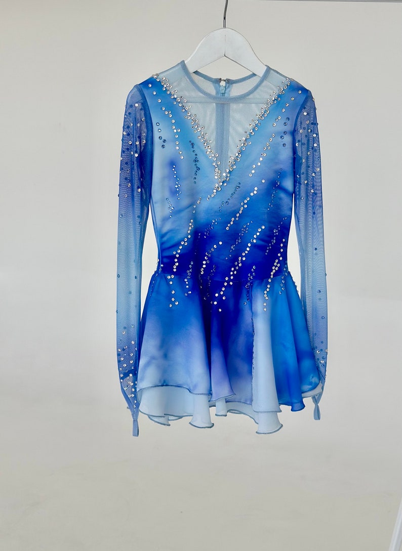 K&ouml;nnte beinhalten: Ein blaues und wei&szlig;es Eiskunstlaufkleid mit langen &Auml;rmeln und einem V-Ausschnitt. Das Kleid ist mit Strasssteinen verziert und hat einen mehrlagigen Rock.