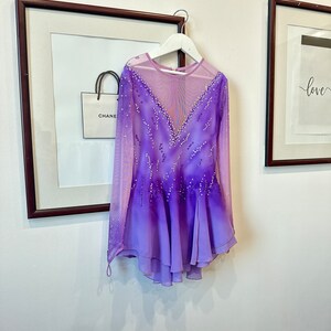 May include: A purple and pink ombre figure skating dress with long sleeves and a sheer bodice. The dress is decorated with rhinestones and has a layered skirt.