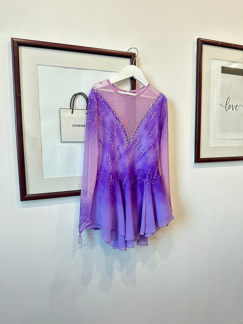 May include: A purple and pink ombre figure skating dress with long sleeves and a sheer bodice. The dress is decorated with rhinestones and has a layered skirt.