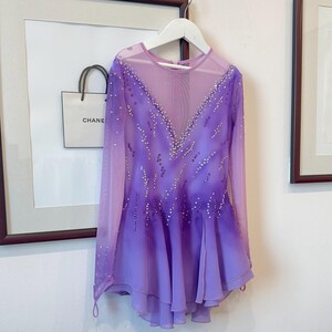 May include: A purple figure skating dress with long sleeves and a gradient design. The dress is decorated with rhinestones and has a sheer bodice.