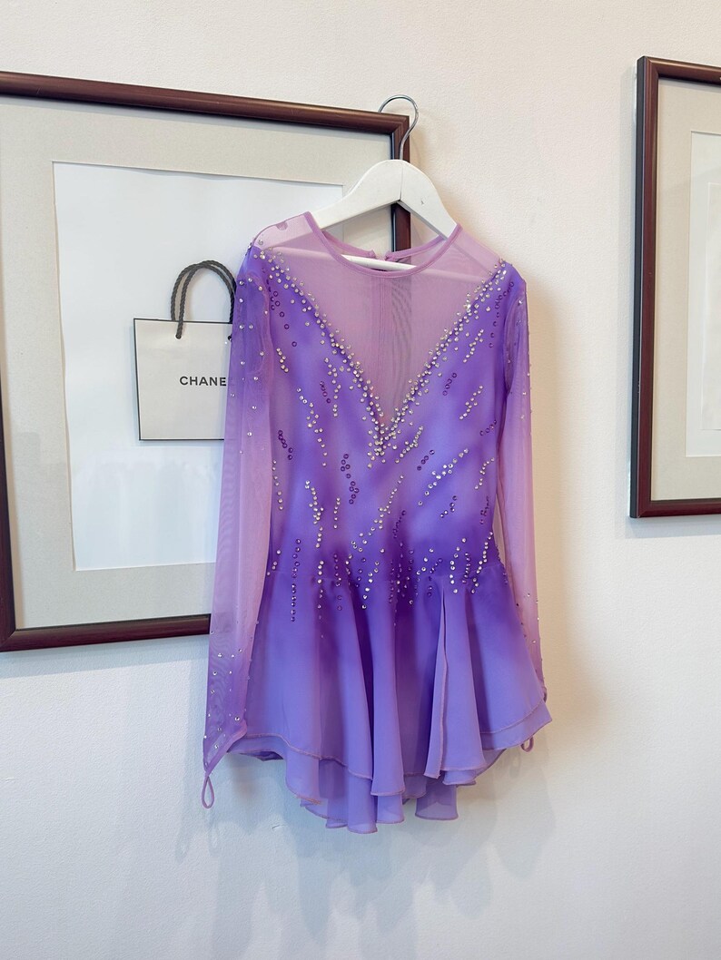 May include: A purple figure skating dress with long sleeves and a gradient design. The dress is decorated with rhinestones and has a sheer bodice.