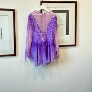 May include: A purple and pink figure skating dress with long sleeves and a mesh back. The dress is decorated with rhinestones.