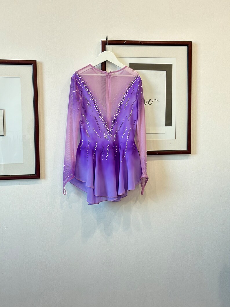 May include: A purple and pink figure skating dress with long sleeves and a mesh back. The dress is decorated with rhinestones.