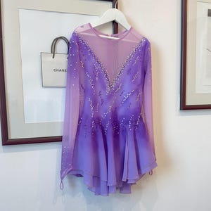 May include: A purple figure skating dress with long sleeves and a sheer bodice. The dress is decorated with rhinestones and has a layered skirt.