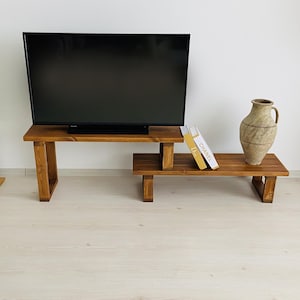 K&ouml;nnte beinhalten: Ein TV-St&auml;nder aus Holz mit zwei Regalen. Das obere Regal h&auml;lt einen Flachbildfernseher und das untere Regal eine Vase und zwei B&uuml;cher. Der St&auml;nder ist aus dunkelbraunem Holz und hat einen rustikalen Look.