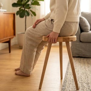 Könnte beinhalten: Ein kleiner, runder Holzstuhl mit drei angewinkelten Beinen. Der Hocker ist aus hellem Holz gefertigt und hat ein schlichtes, minimalistisches Design. Der Hocker ist etwa 30 cm hoch und eignet sich zum Sitzen oder als dekoratives Element.