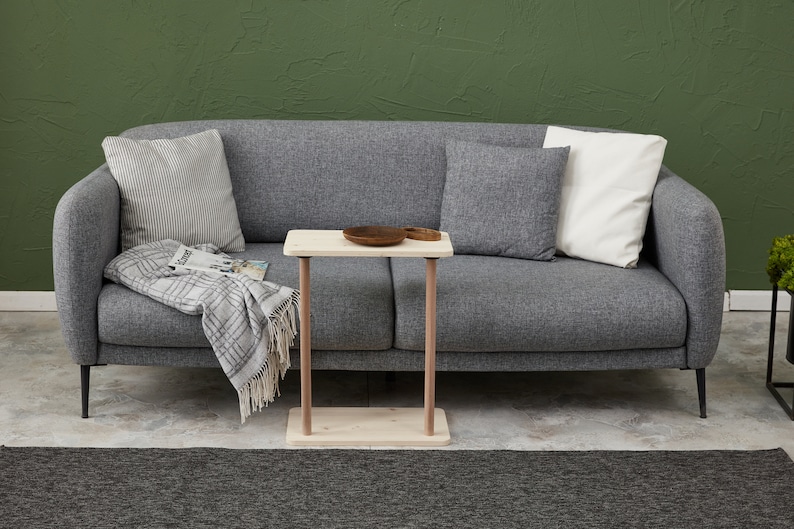 Może przedstawiać: Jasnoszara sofa z białym kocem i poduszkami. Mały drewniany stolik z drewnianą miską stoi przed sofą.