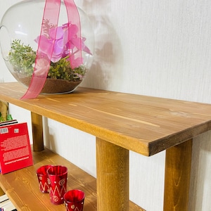 May include: A wooden shelf unit with two shelves. The top shelf has a glass globe with pink flowers and greenery inside. The bottom shelf has a stack of books and three red candle holders.
